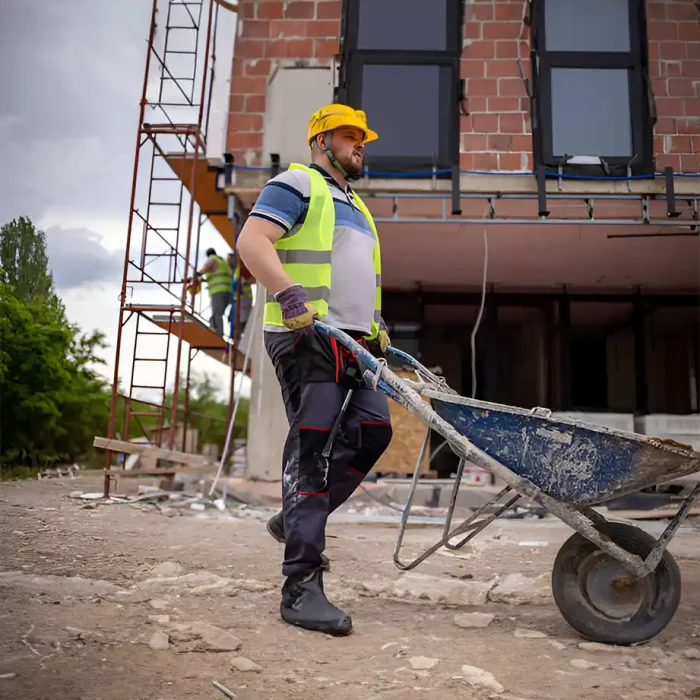 Arbeiter mit gelbem Schutzhelm und Warnweste, der auf einer Baustelle eine Schubkarre schiebt in Sinsheim