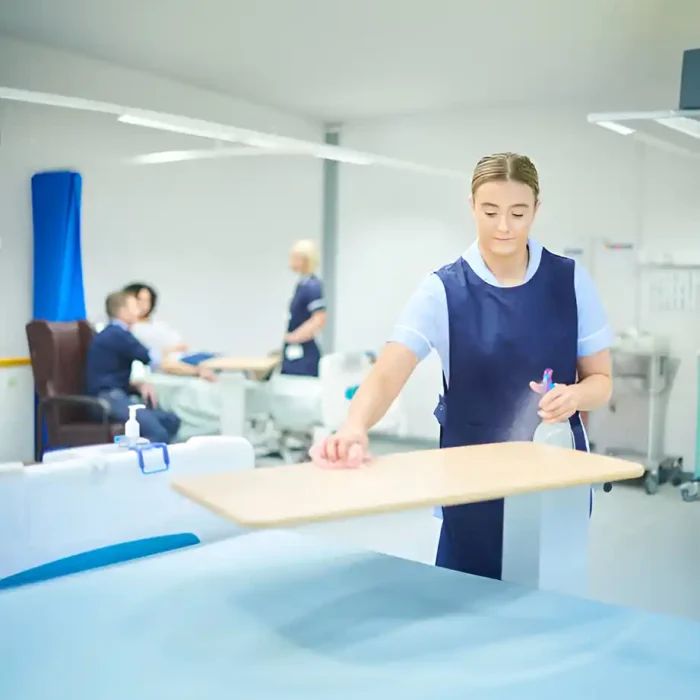 Mitarbeiter des Gesundheitswesens reinigen einen Nachttisch mit einer Sprühflasche in einer geschäftigen Krankenhausumgebung in Friedberg