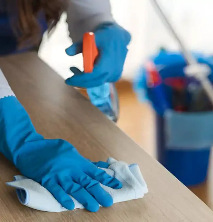 Person mit blauen Handschuhen, die einen Holztisch mit einem Tuch und einer Sprühflasche abwischt