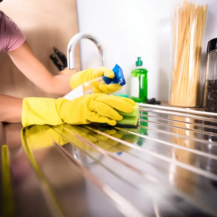 Person mit gelben Handschuhen reinigt eine Edelstahlspüle mit einem Schwamm und einer Sprühflasche in Coburg