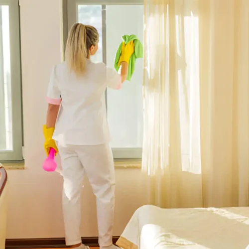 Frau in weißer Uniform putzt ein Fenster mit gelben Handschuhen und einem grünen Tuch