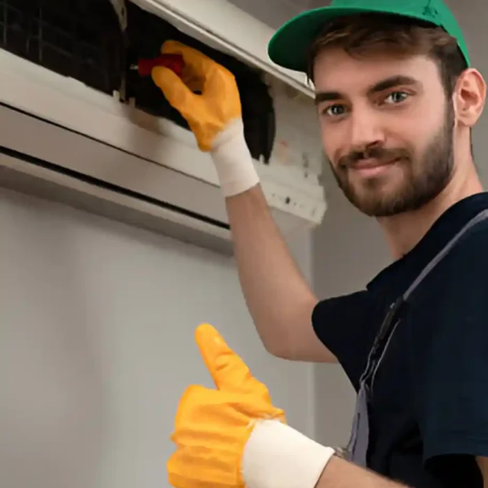 Lächelnder Techniker mit grüner Mütze und orangefarbenen Handschuhen bei der Wartung einer Klimaanlage in Coburg