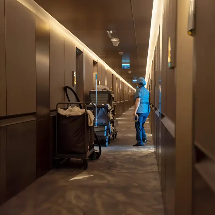 Ein Hotelreiniger in Uniform, der einen Wagen durch einen gut beleuchteten Flur mit braunen Wänden schiebt