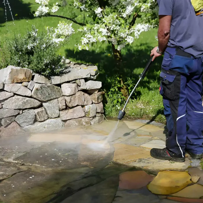 Ein Arbeiter wäscht mit Hochdruck eine Steinterrasse in der Nähe einer Felswand und eines blühenden Baums