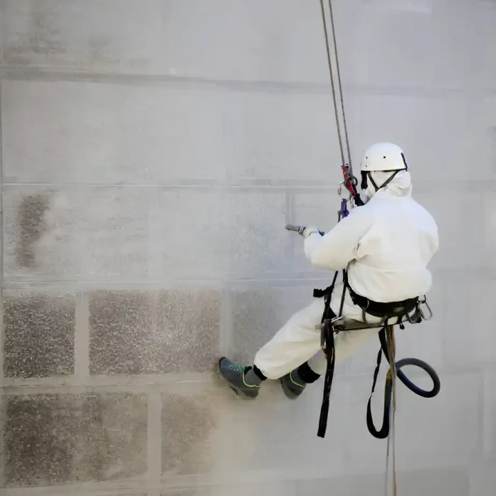 Arbeiter in Schutzausrüstung benutzt Seilzugang, um eine Gebäudewand mit Druck zu reinigen