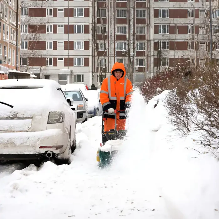 Ein Arbeiter in orangefarbener Kleidung benutzt eine Schneefräse auf einem verschneiten Gehweg in der Stadt