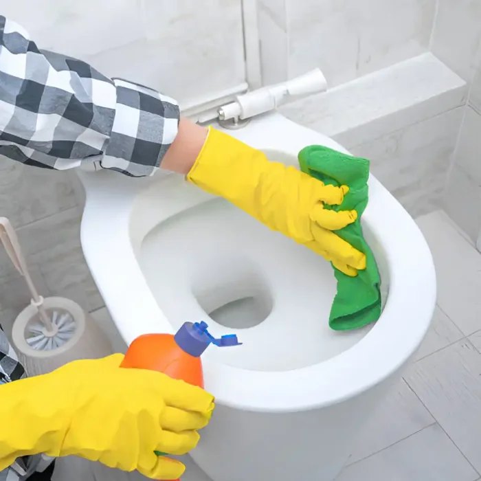 Person mit gelben Handschuhen, die in einer Hand ein Reinigungsmittel und einen Lappen hält, um den Toilettensitz zu reinigen